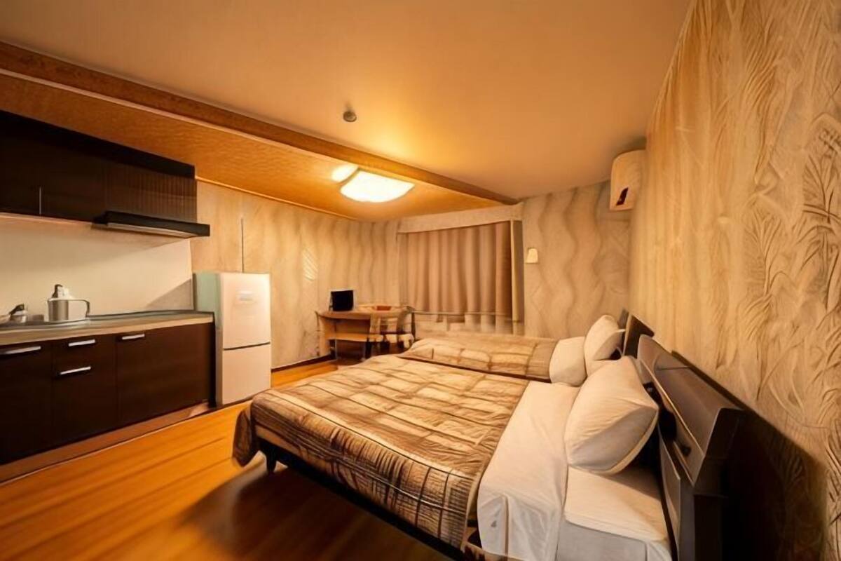 A compact hotel room with a kitchenette, two beds, a small table, and a patterned wall.