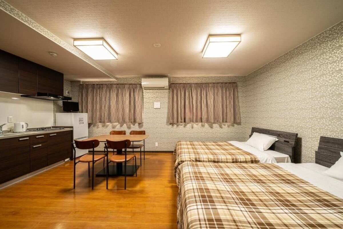A compact hotel room with a kitchenette, a dining area with a table and chairs, two beds with plaid bedding, and a textured wall.