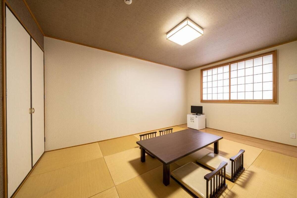 A traditional Japanese room with tatami flooring, a low table, benches, a sliding door, a window with shoji screens, and a ceiling light fixture.
