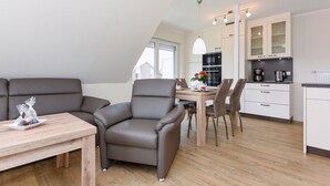 A modern living room with a sofa, armchair, and dining table. A kitchen area with cabinets and appliances is visible in the background. - Vacation apartment top right - Haus Fallen Anker in Neuharlingersiel (Neuharlingersiel)