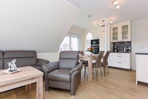 Uma sala de estar moderna com sofá, poltrona e mesa de jantar. Uma cozinha com armários e eletrodomésticos é visível ao fundo.