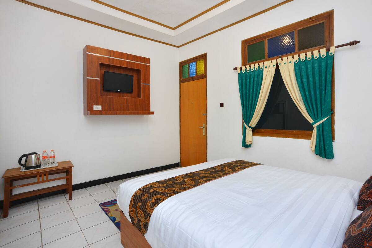 Una habitación de hotel con una cama, un televisor montado en la pared, una puerta de madera y una ventana con cortinas.