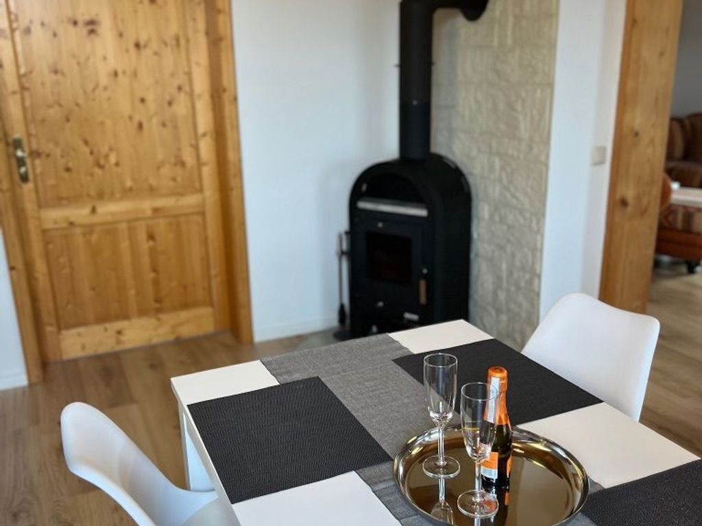 Une table à manger recouverte d’une nappe à carreaux, avec deux verres à vin et une bouteille de vin. On aperçoit un poêle à bois noir à l’arrière-plan.