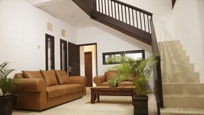 Un salon avec un canapé en cuir marron, une table basse en bois et deux plantes en pot. Un escalier avec une rampe en bois est visible sur la droite.