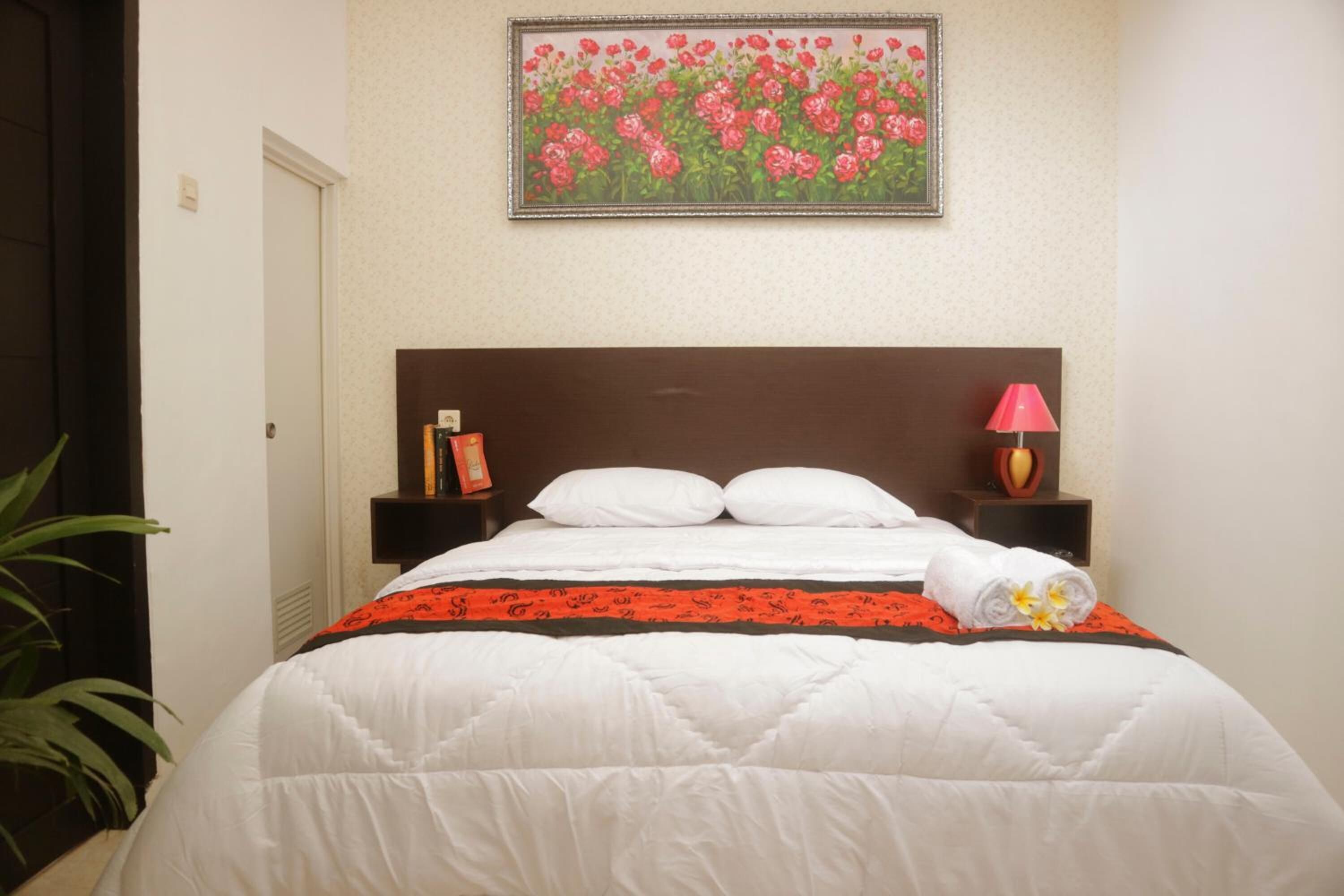 A neatly made bed with white and red bedding, a framed floral artwork above it, and a bedside lamp.