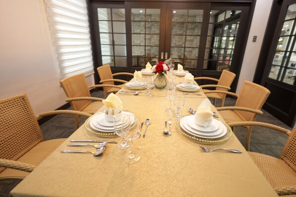 Una mesa de comedor con platos blancos, copas con detalles dorados y cubiertos, rodeada de sillas de mimbre.