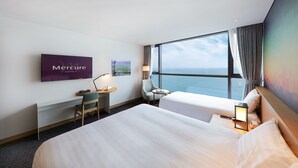 Habitación de hotel con un ventanal grande que ofrece vistas al mar, un escritorio con silla, dos camas y un televisor de pantalla plana en la pared.