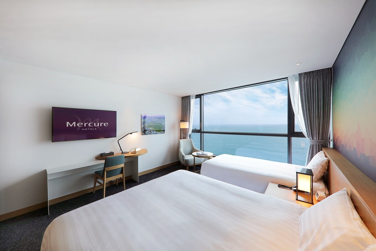 A hotel room with a large window offering a view of the sea, a desk with a chair, two beds, and a flat-screen TV on the wall.