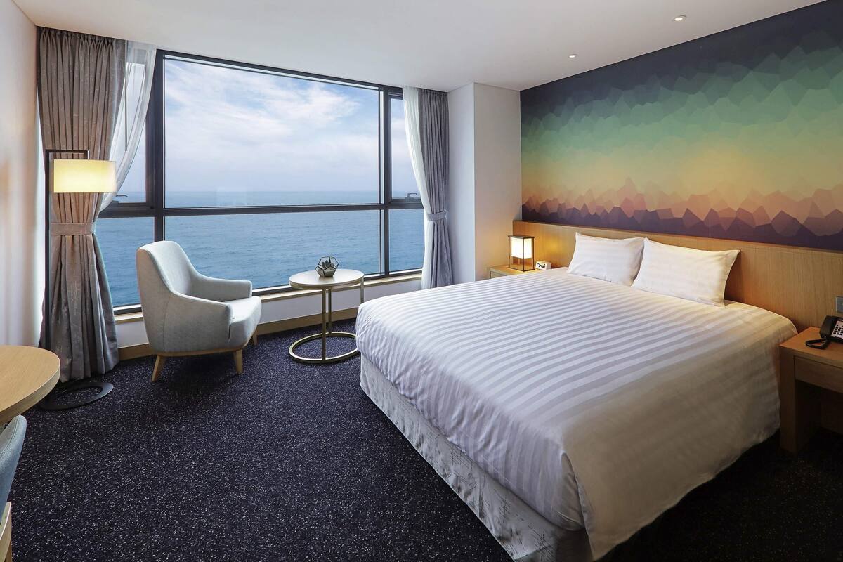 A hotel room with a large bed, a chair, a small table, and a view of the sea.