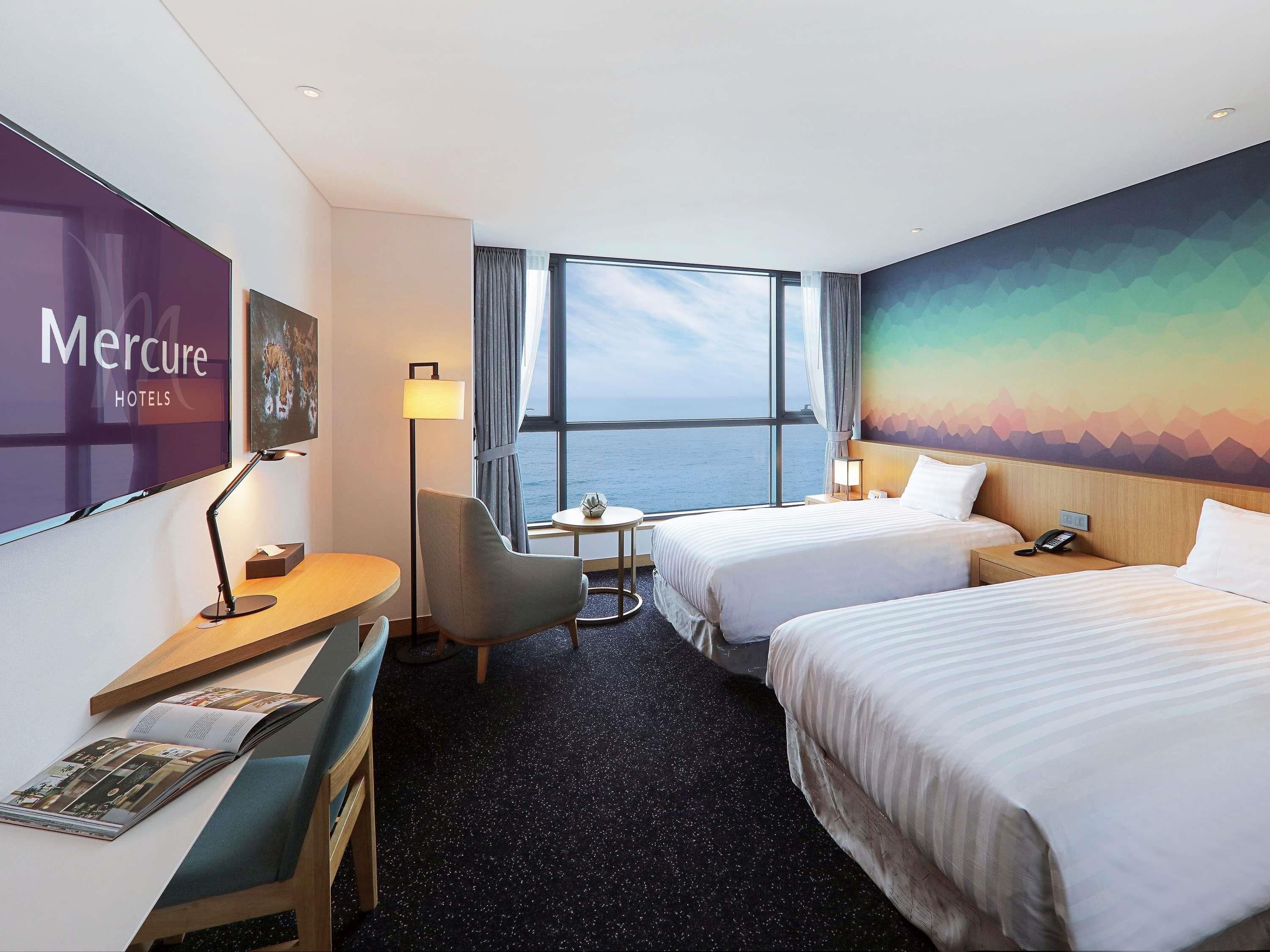 Ein Hotelzimmer mit zwei Betten, einem Schreibtisch, einem Sessel, einem Fernseher und einem großen Fenster mit Meerblick.