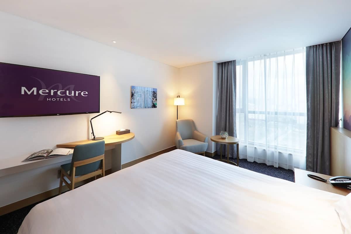A hotel room with a bed, a desk with a lamp, a chair, a small table, and a TV displaying the hotel name.