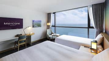A hotel room with a bed, a desk, a chair, a TV, and a window with a view of the sea.
