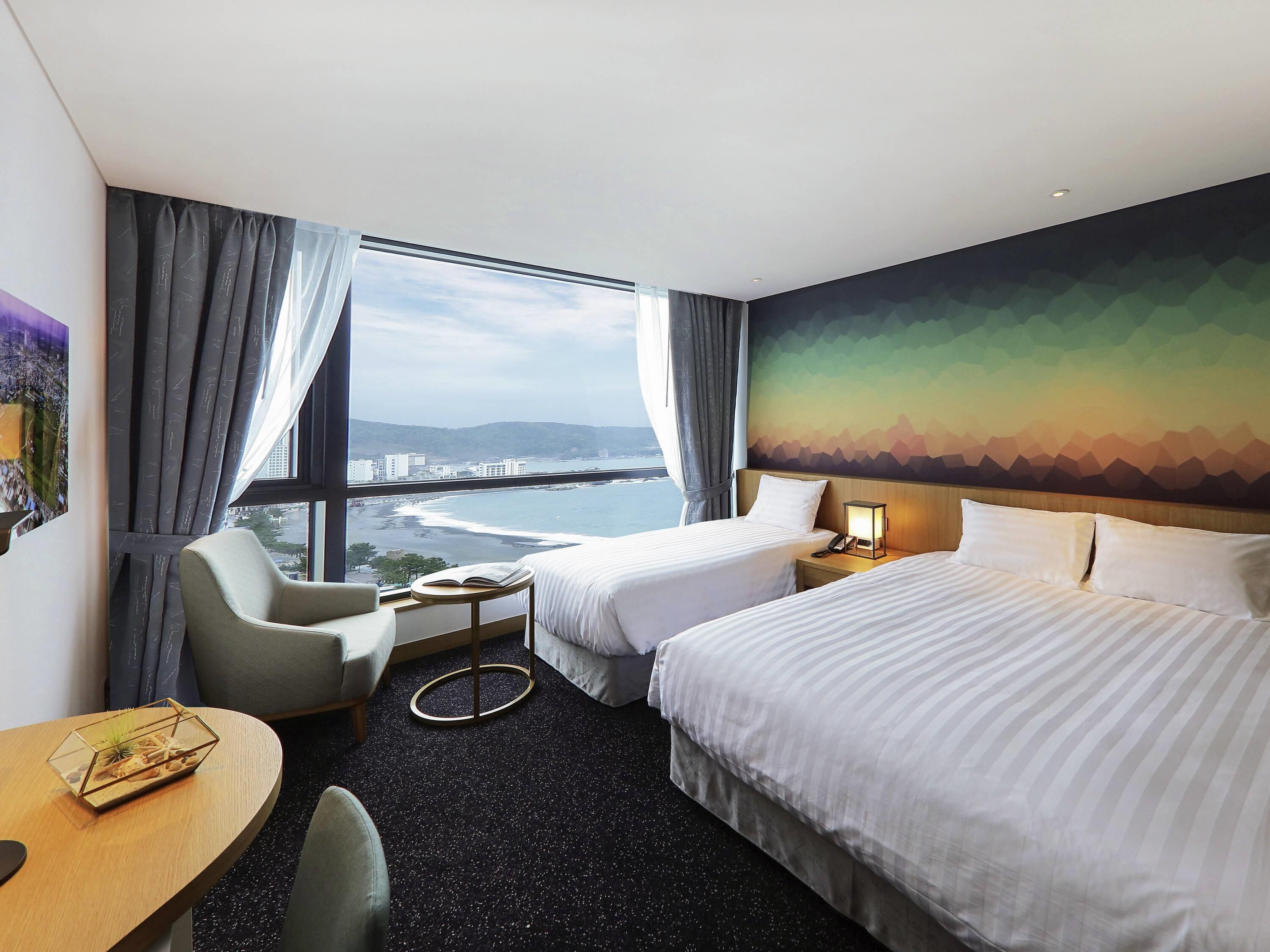 A hotel room with two beds, a desk, a chair, a small table with a book, and a view of a coastal area.