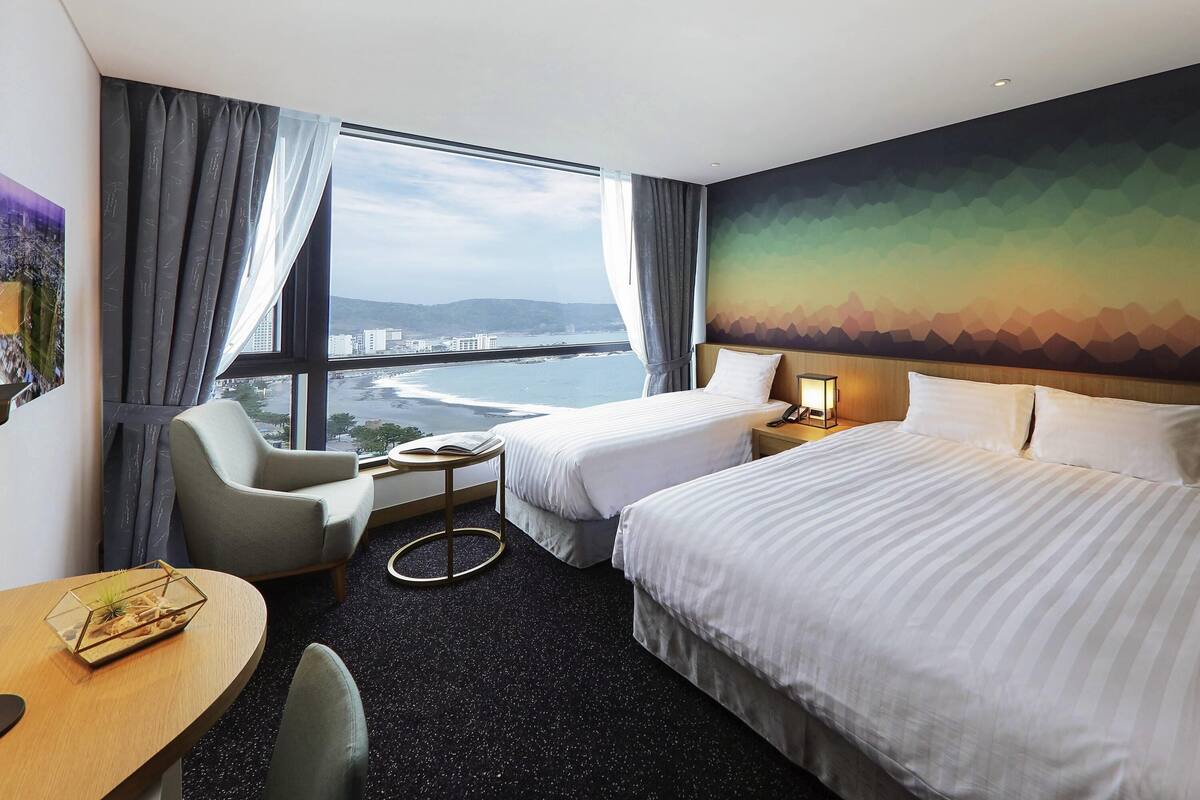 A hotel room with two beds, a desk, a chair, a small table with a book, and a view of a coastal area.