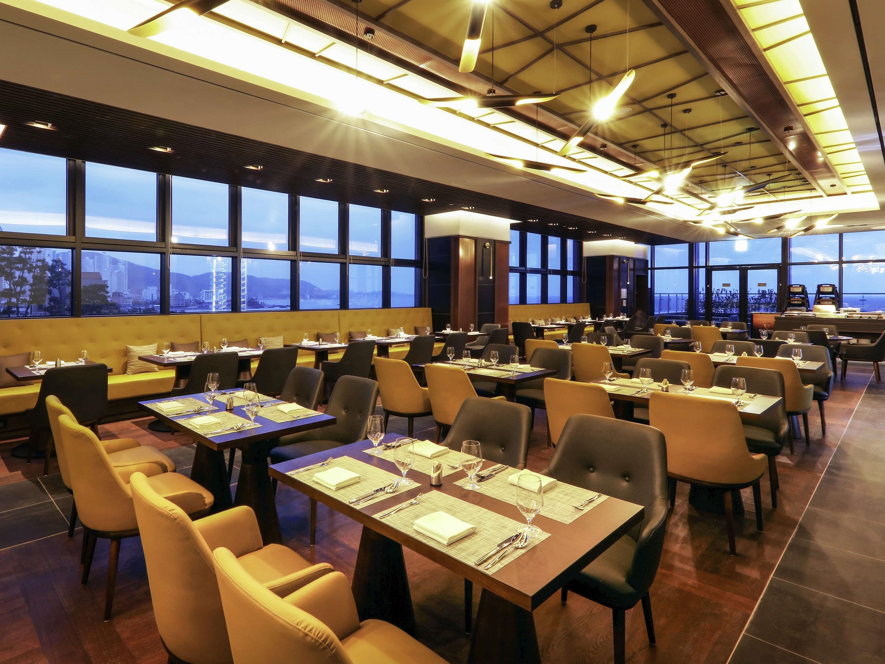 A spacious restaurant with multiple dining tables and chairs, large windows offering a city view, and modern lighting fixtures.