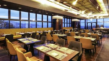 A spacious restaurant with multiple dining tables and chairs, large windows offering a city view, and modern lighting fixtures.