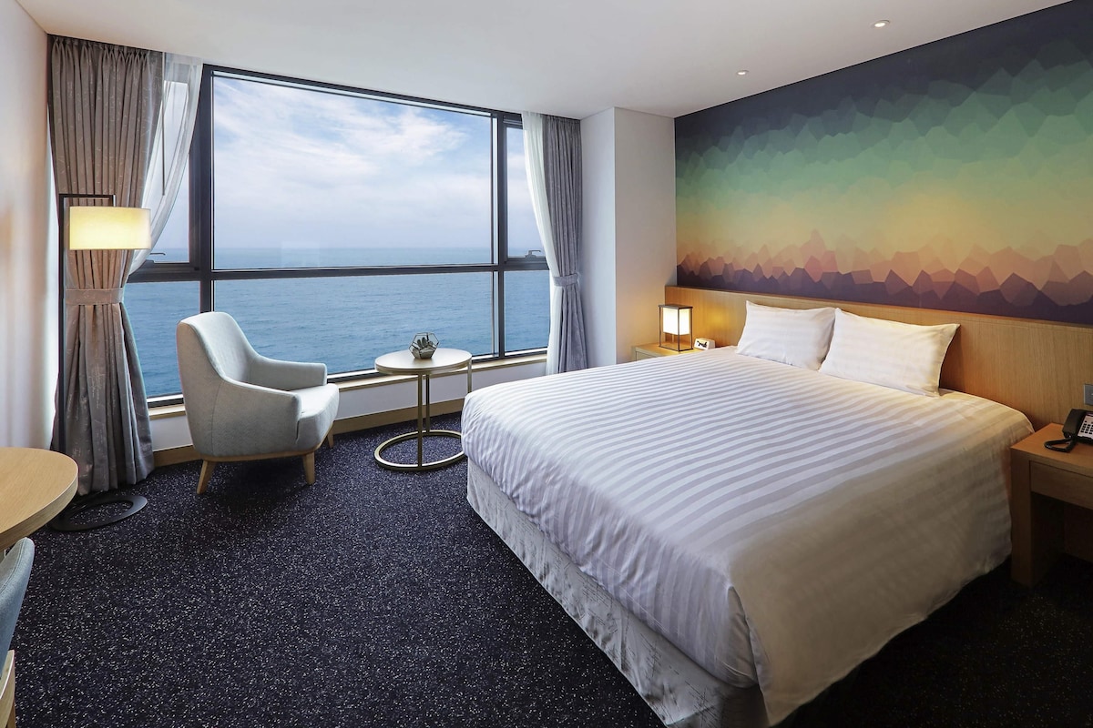 A hotel room with a large bed, a chair, a small table, and a view of the sea.