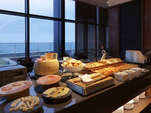 A buffet with various cheeses, breads, and pastries, with a view of the ocean through large windows. - Mercure Ambassador Ulsan (Ulsan)