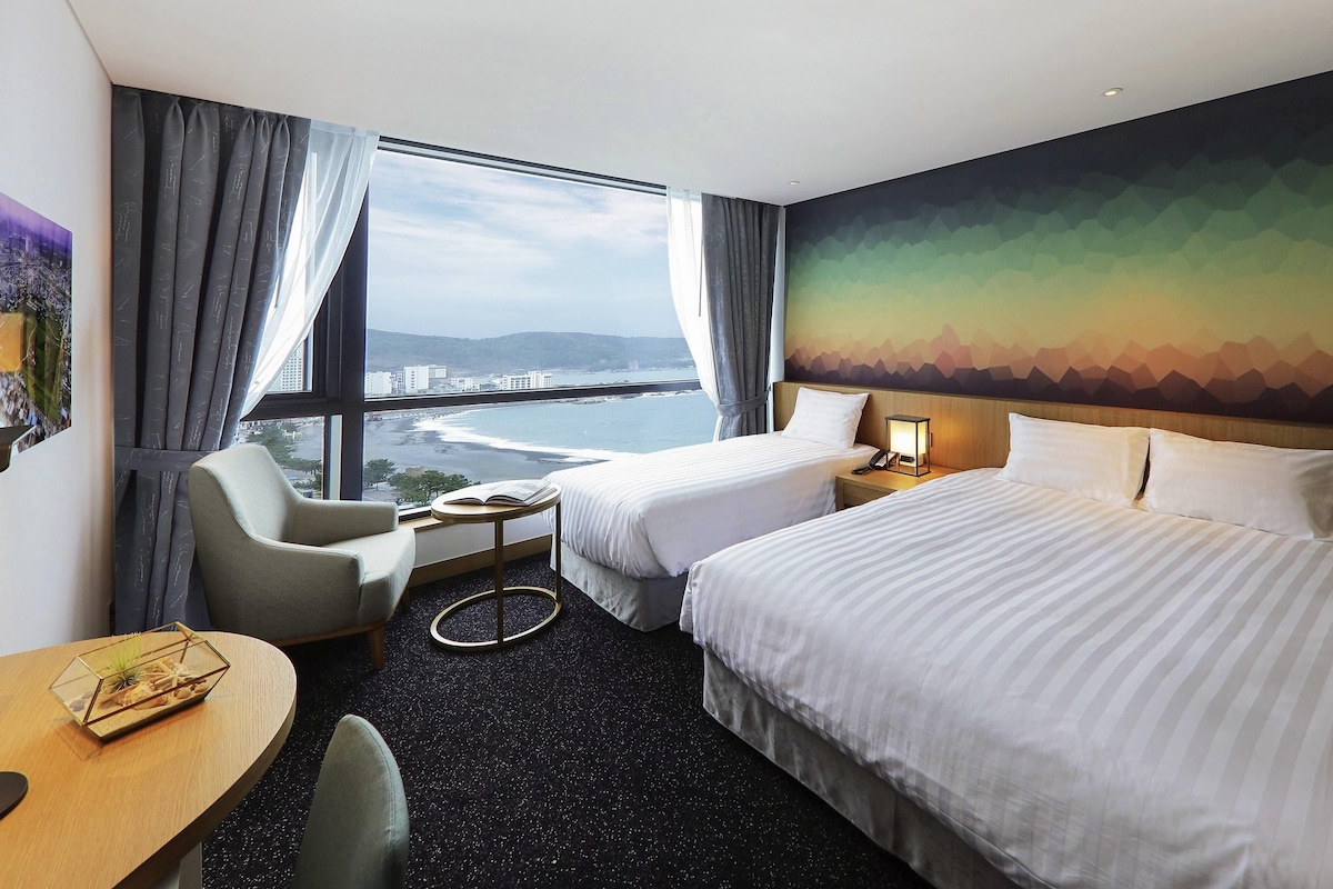 A hotel room with two beds, a desk, a chair, a small table with a book, and a view of a coastal area.