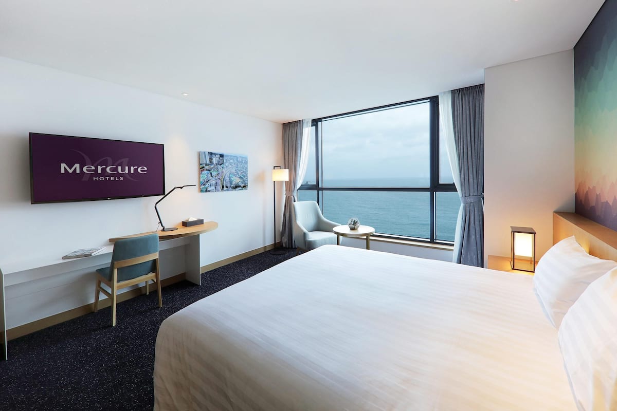 Habitación de hotel con cama, escritorio, silla, televisión, ventana con vista al mar y un cuadro en la pared.