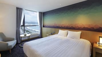 Une chambre d’hôtel avec un grand lit, une chaise, une petite table, une table de chevet et une vue sur la plage par la fenêtre.