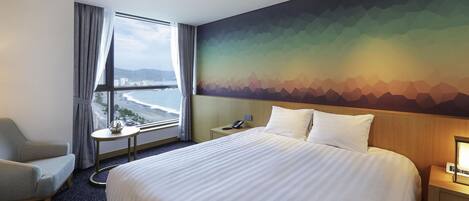 A hotel room with a large bed, a chair, a small table, a nightstand, and a view of the beach through a window.