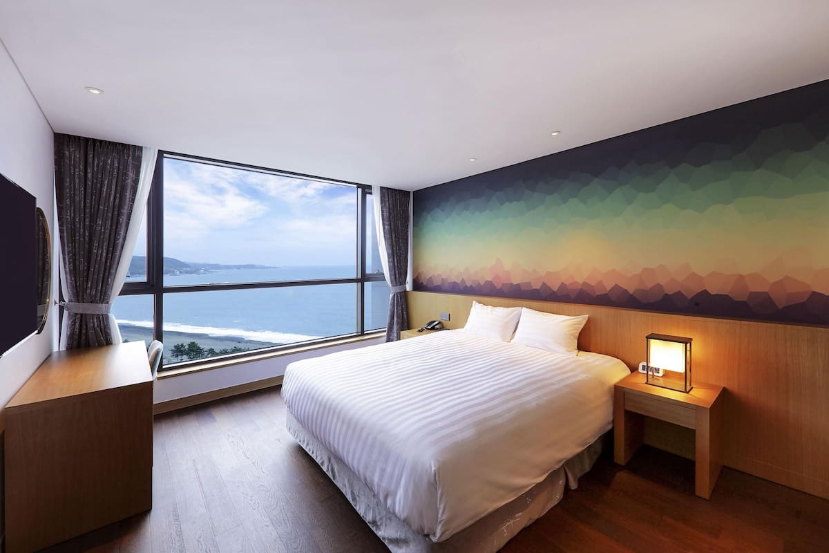 A hotel room with a large window, a bed, a bedside table with a lamp, and a wall mural.