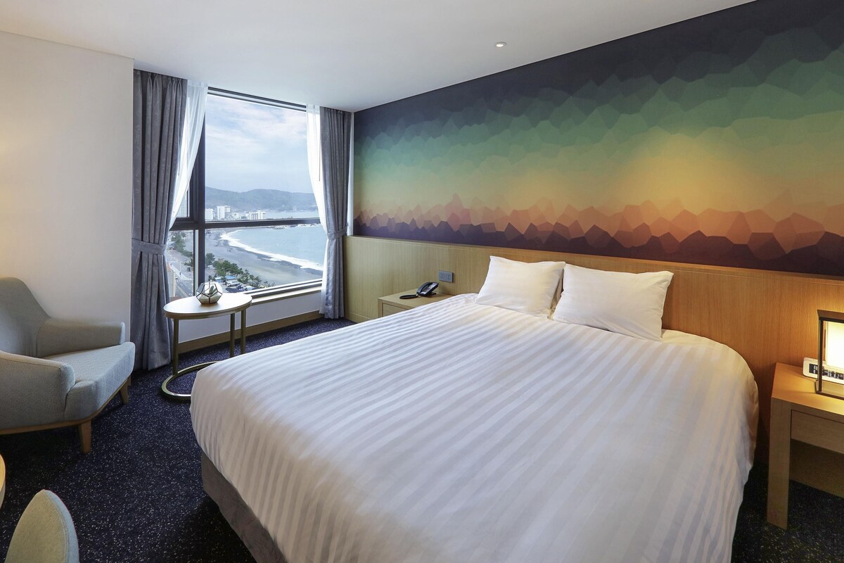 A hotel room with a large bed, a chair, a small table, a nightstand, and a view of the beach through a window.