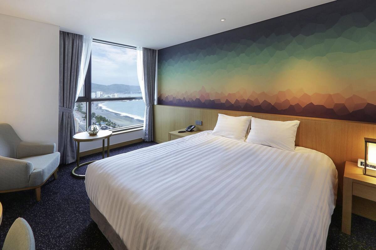 A hotel room with a large bed, a chair, a small table, a nightstand, and a view of the beach through a window.