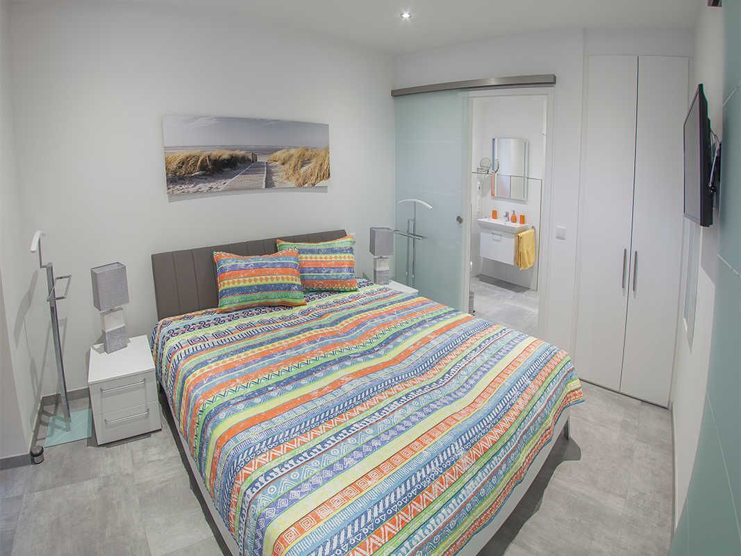 A modern bedroom with a large bed, a bedside table, a nightstand, a glass shower enclosure, a wardrobe, and a mounted TV.