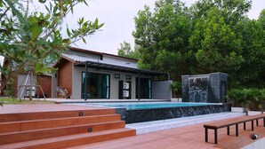Un espace piscine extérieur moderne, agrémenté d’un escalier en bois menant à la piscine, d’une cascade en pierre et d’un bâtiment doté de grandes fenêtres et d’un patio couvert.