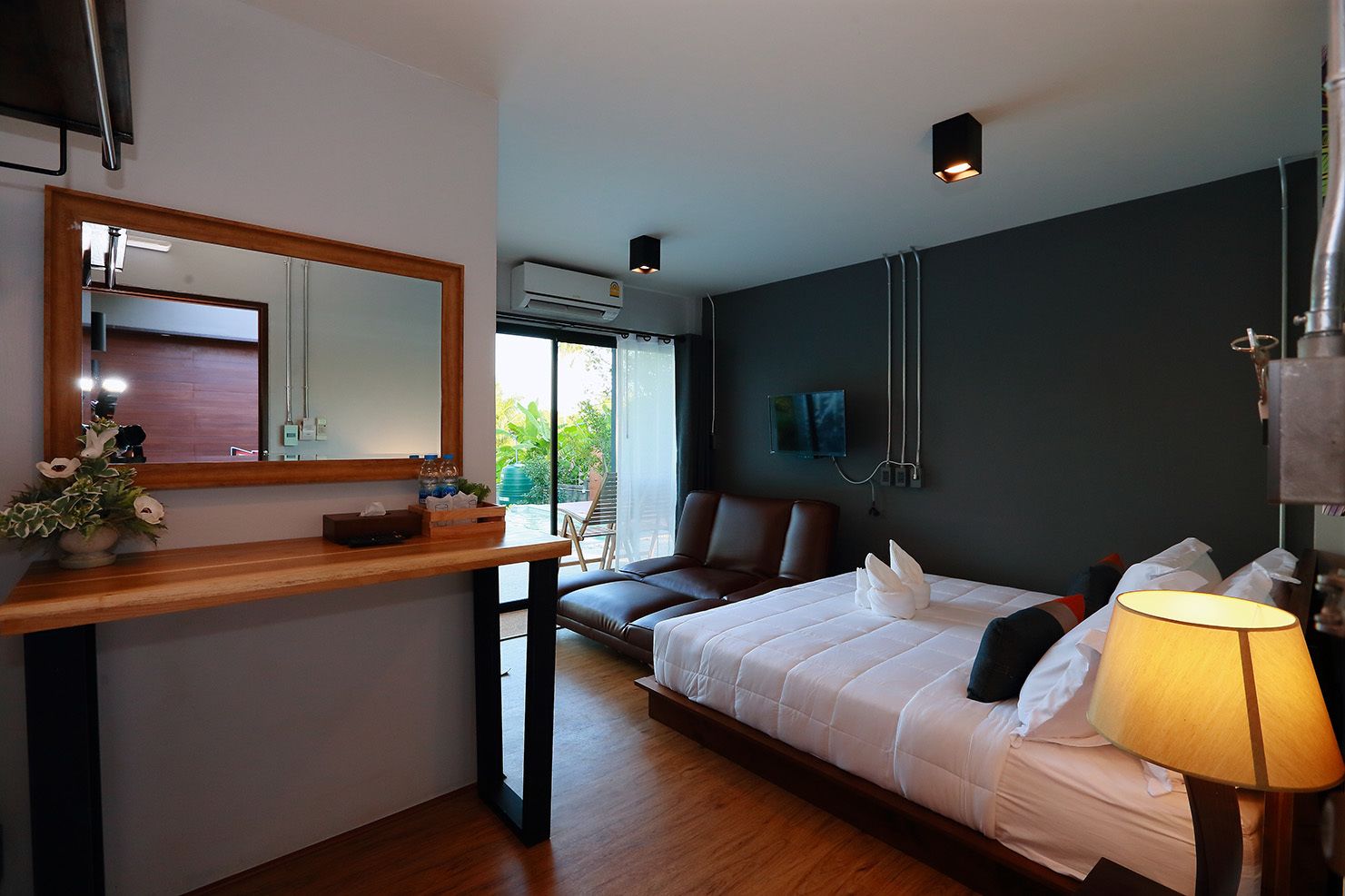 A modern hotel room with a wooden desk, a large bed, a bedside lamp, a TV, and an air conditioning unit.