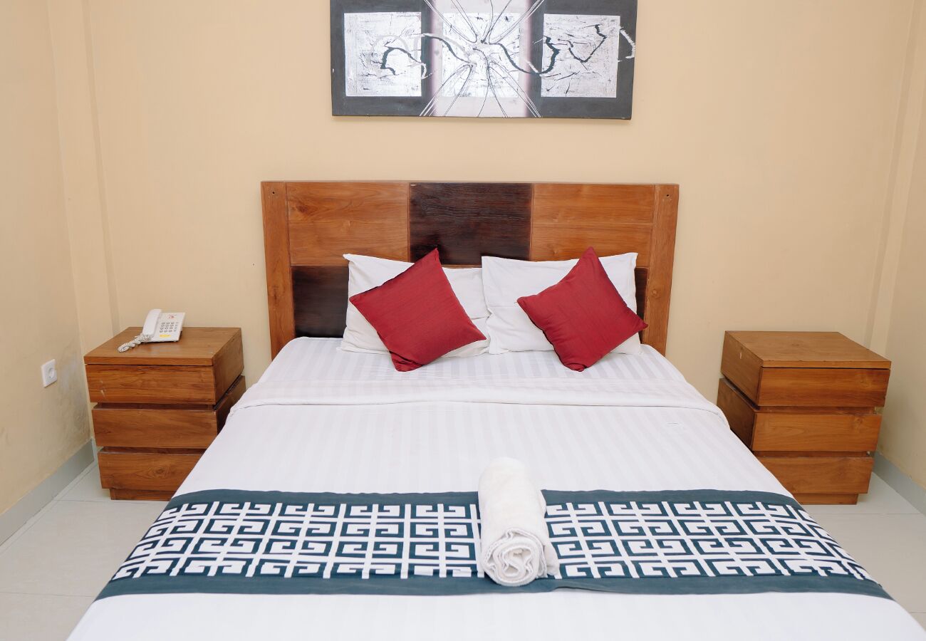 A neatly made bed with a patterned bedspread, two red pillows, and a rolled towel on the floor. There are two wooden nightstands with a phone on one.