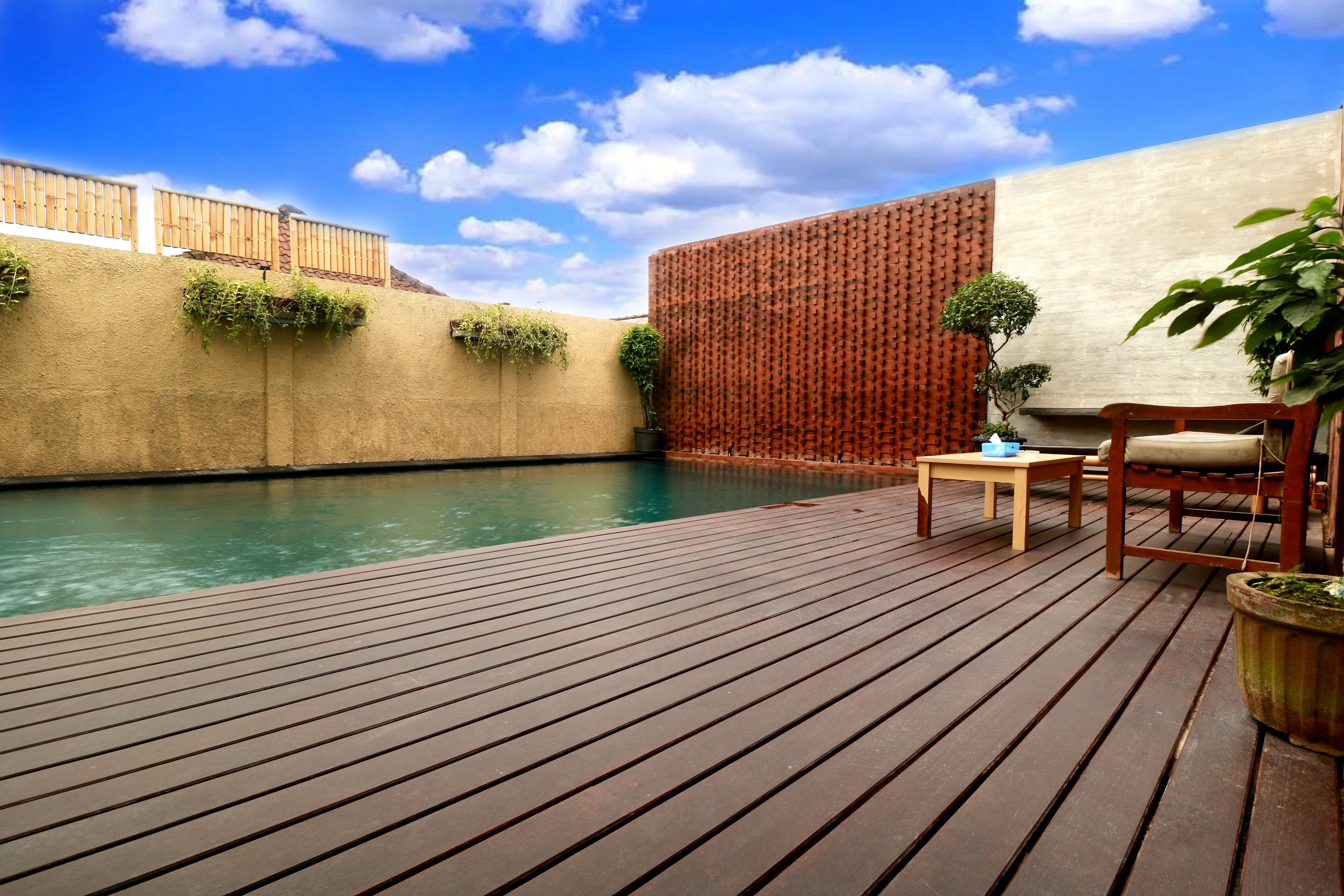 Una terraza de madera con piscina, una silla de madera, una mesita y plantas en macetas.