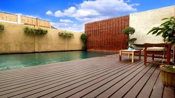 Una terraza de madera con piscina, una silla de madera, una mesita y plantas en macetas.