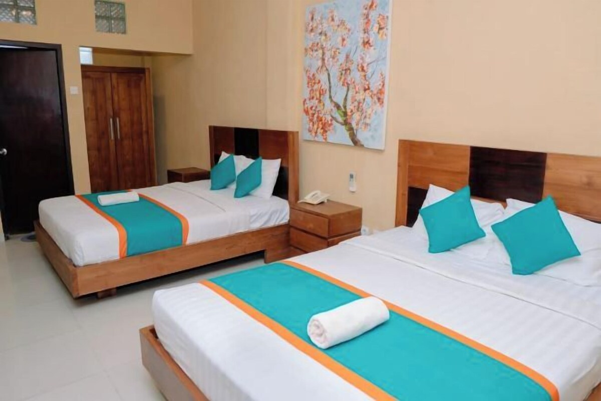 Une chambre d’hôtel avec deux lits, chacun doté d’une tête de lit en bois, de draps blancs et d’éléments de décoration de couleur turquoise. Un tableau orne le mur et une armoire en bois est présente dans la pièce.
