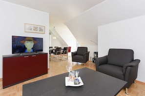 A modern living room with a flat-screen TV, a sofa, a coffee table, and a dining area in the background. - Apartment top left - Haus Addenhausen (Neuharlingersiel)