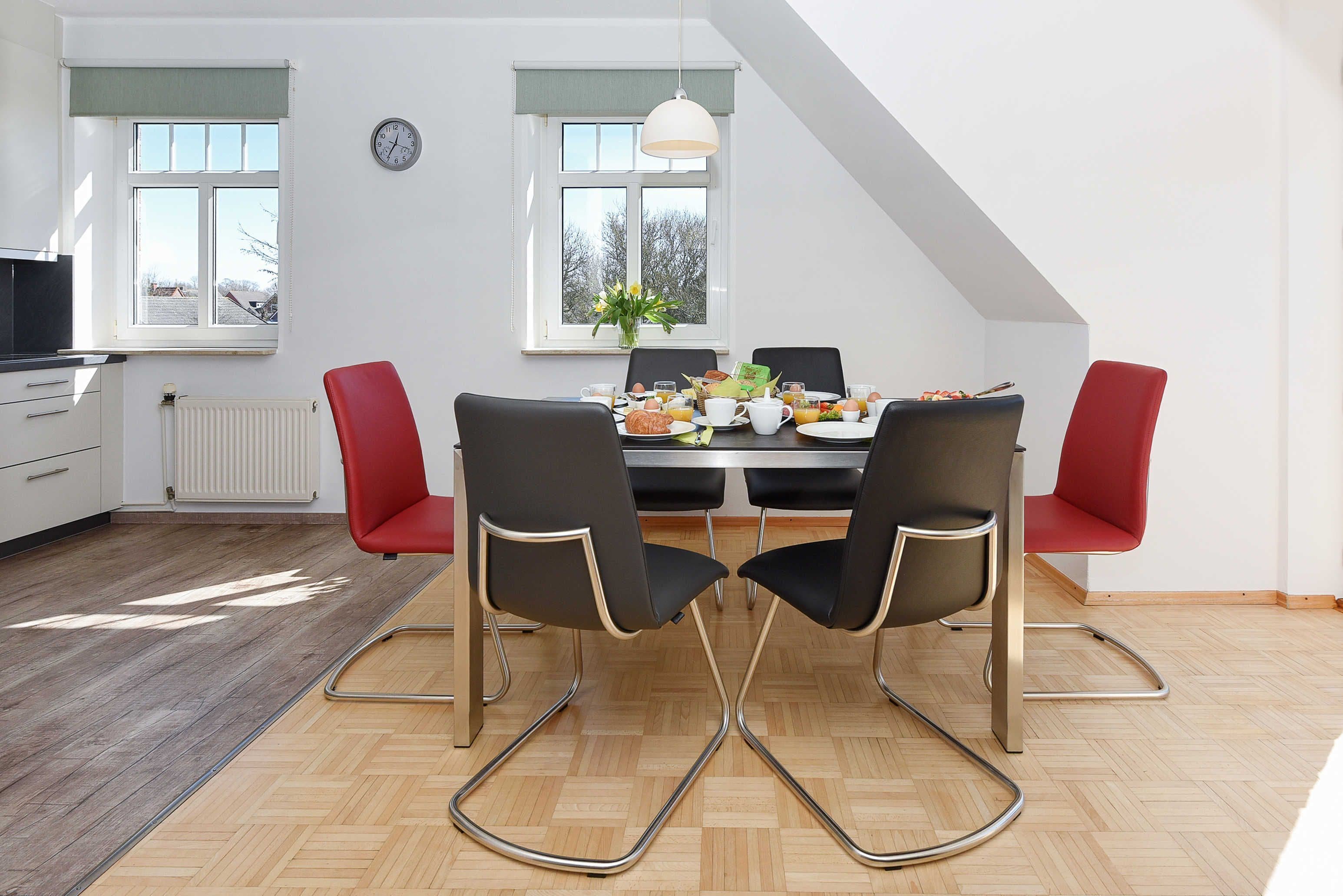 Uma sala de jantar com uma mesa posta para quatro pessoas, duas cadeiras vermelhas e uma cadeira preta com design exclusivo. Há um relógio na parede e uma janela com vista para árvores.