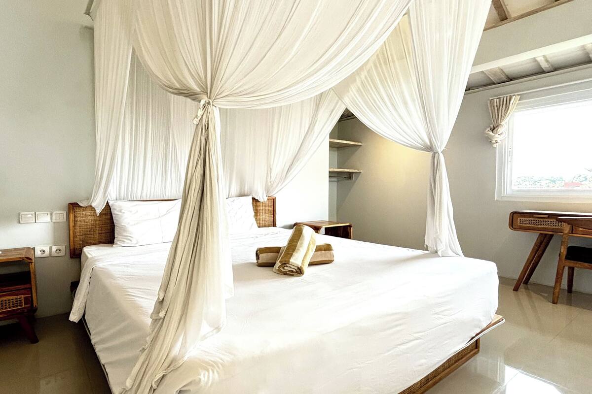 A bedroom with a four-poster bed, a wooden nightstand, a wooden chair, and a window with curtains.