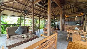 An open-air dining area with wooden furniture, a thatched roof, and a kitchen area in the background.