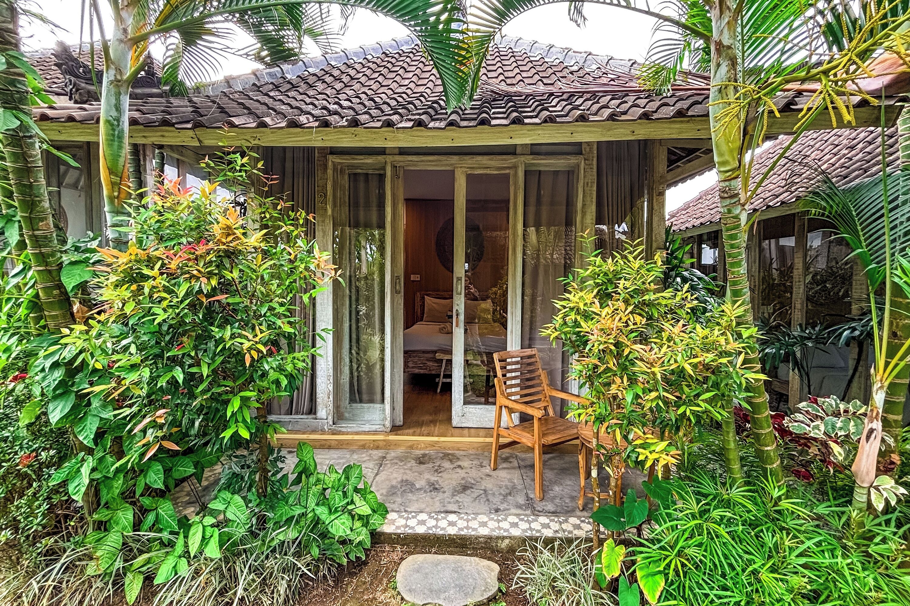 Une maison traditionnelle avec un toit en tuiles, une véranda en bois et une porte-fenêtre coulissante donnant sur une pièce comprenant un lit et une chaise.