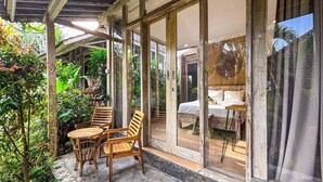 A covered outdoor seating area with wooden chairs and tables, surrounded by lush greenery and large glass doors leading to a bedroom with a bed and a nightstand.