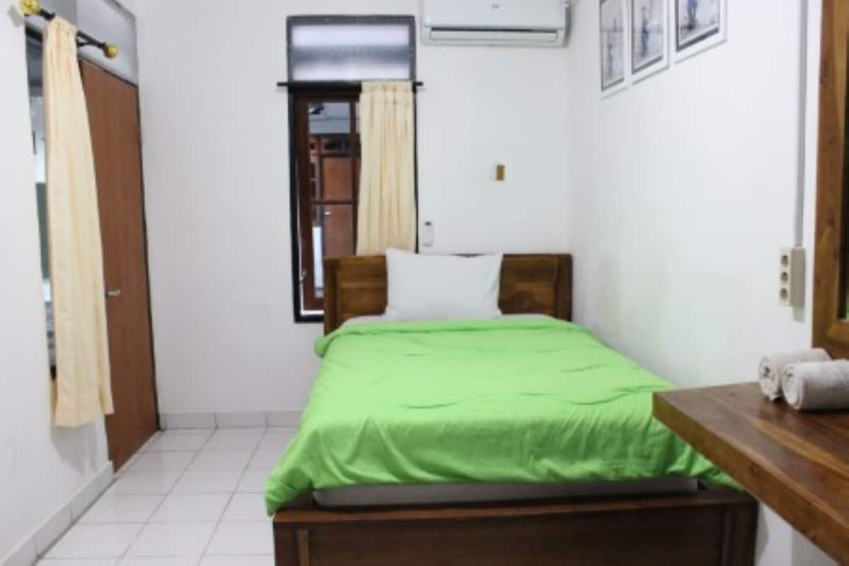 A small, clean room with a single bed, a wooden headboard, a window with curtains, a small table with a towel, and a wall-mounted air conditioner.