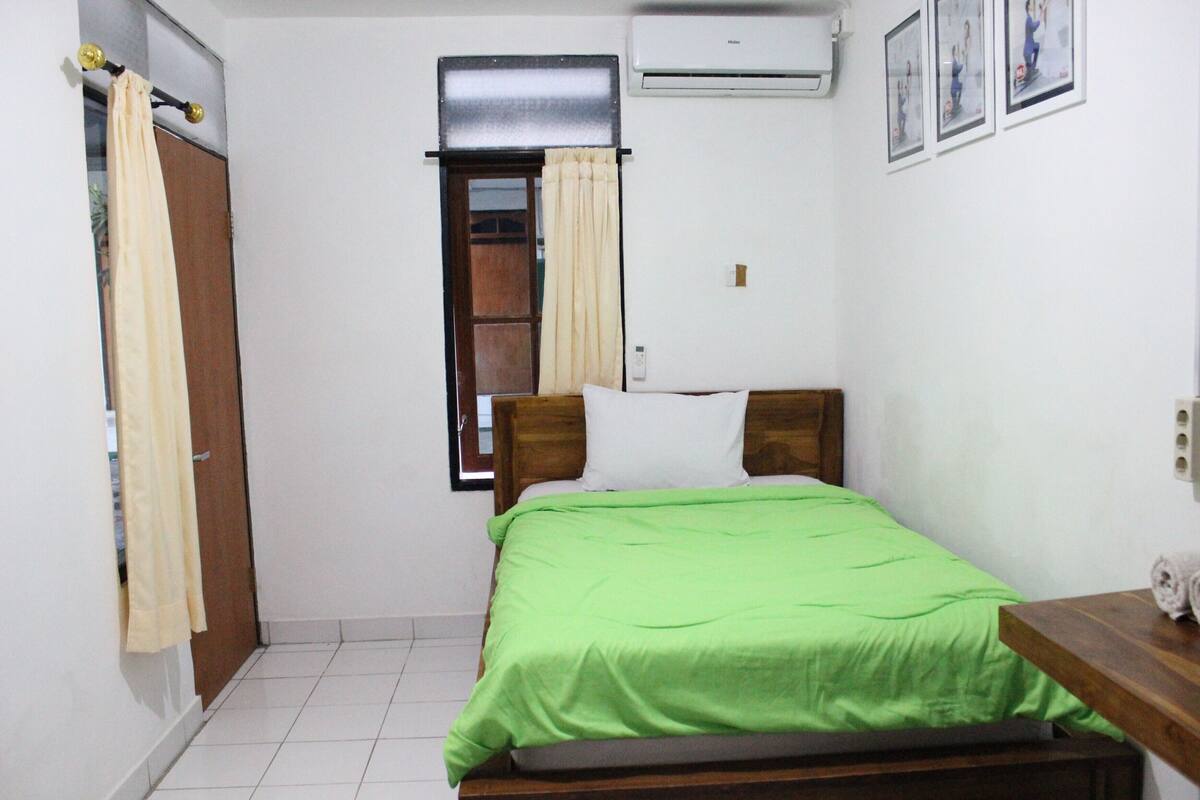 A small, clean room with a bed, a wooden nightstand, a window with curtains, and a wall-mounted air conditioner.