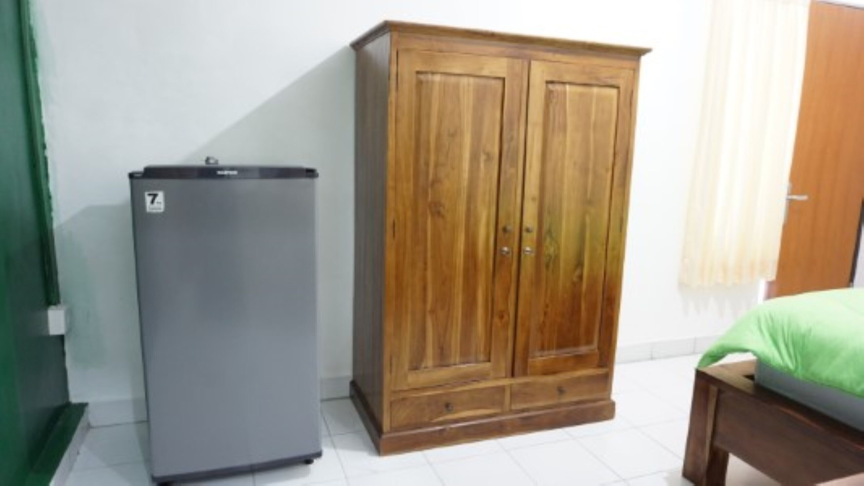 A room with a wooden wardrobe, a grey refrigerator, and a bed with a green cover.
