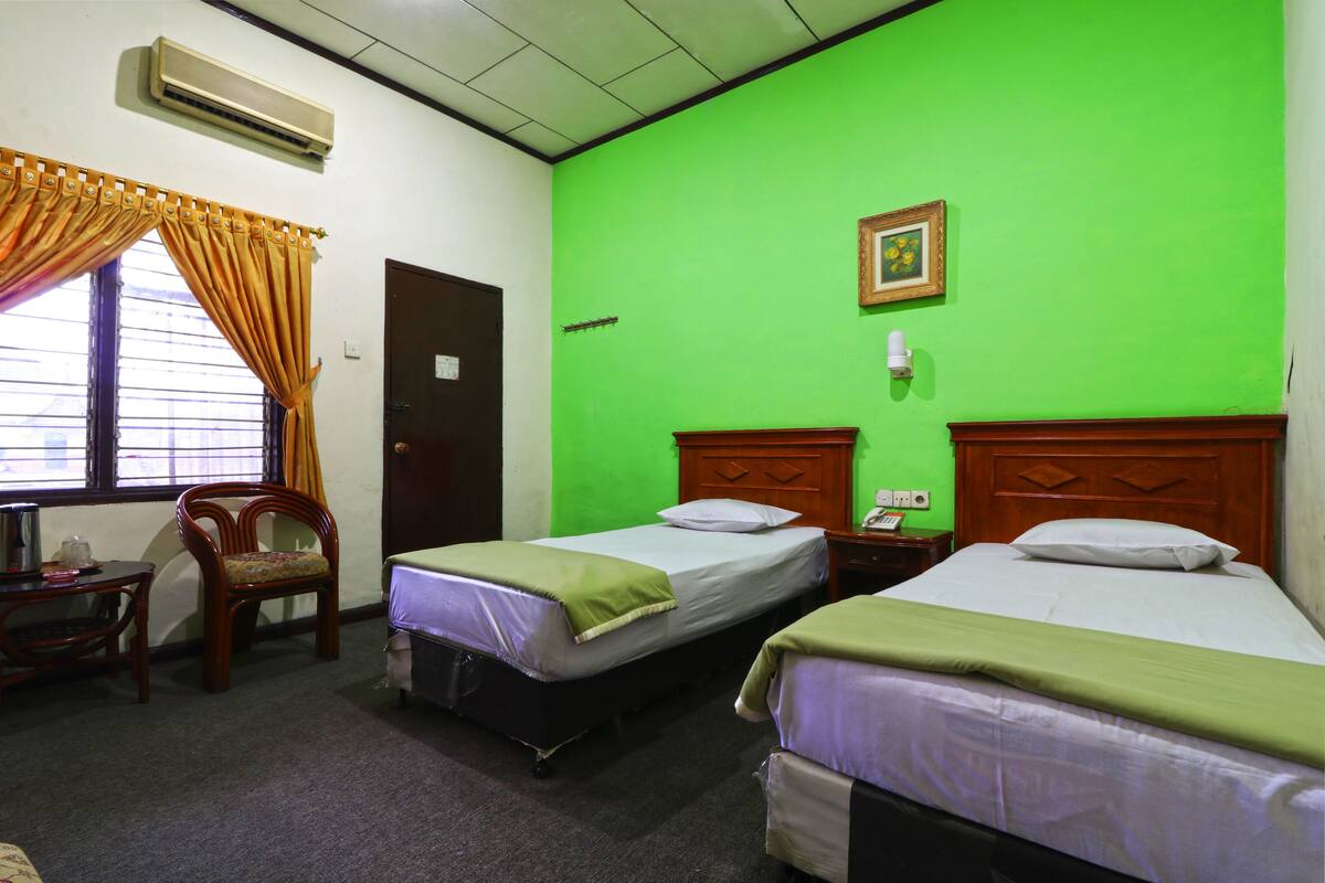 A hotel room with two beds, a wooden chair, a desk with a lamp, and a window with curtains.