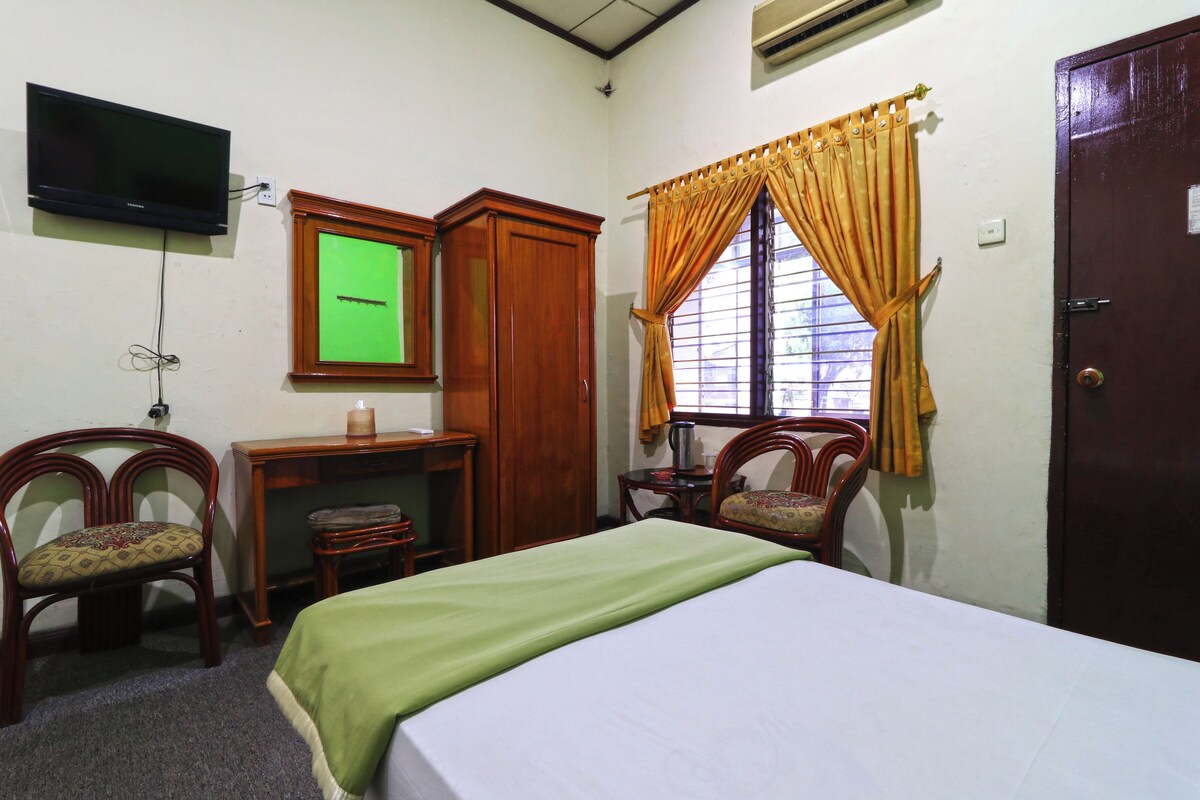 A room with a bed, two chairs, a desk, a television mounted on the wall, and a window with curtains.