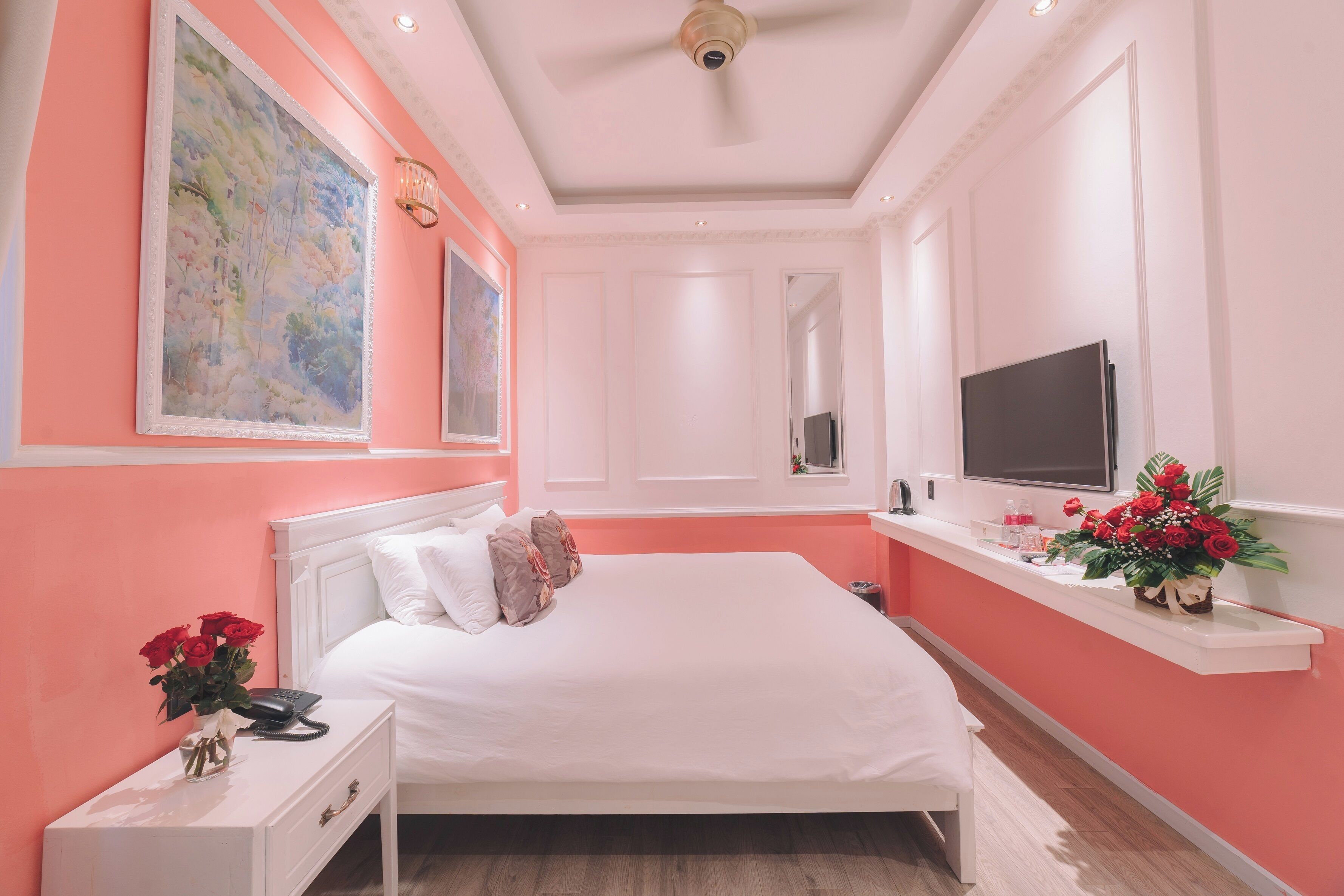 A bedroom with a large bed, a bedside table with a phone and vase of flowers, a TV on a stand, and a painting on the wall.
