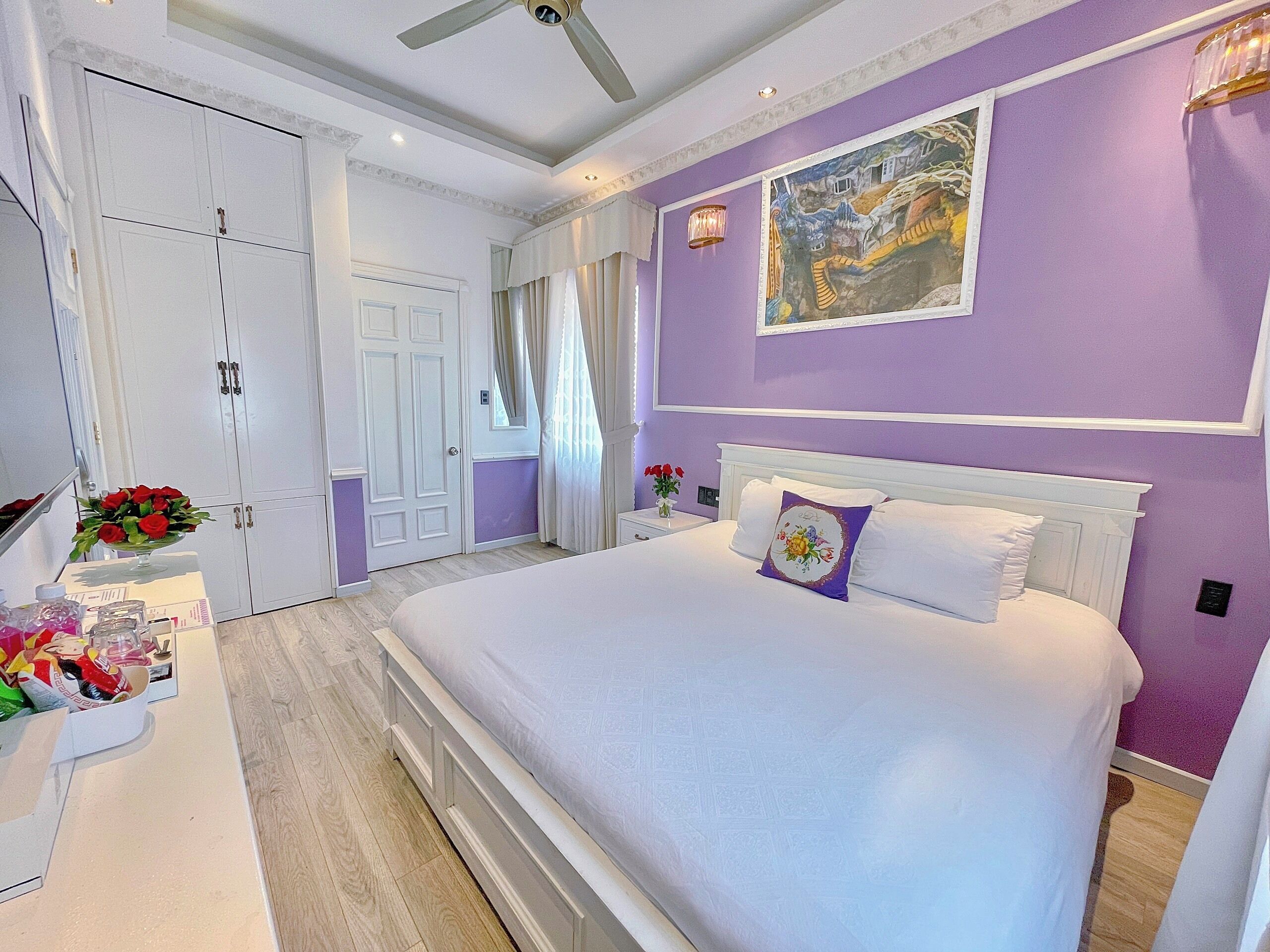 A bedroom with a large bed, white headboard, and a purple accent wall. There is a door with a window, a nightstand with a vase of flowers, and a ceiling fan.