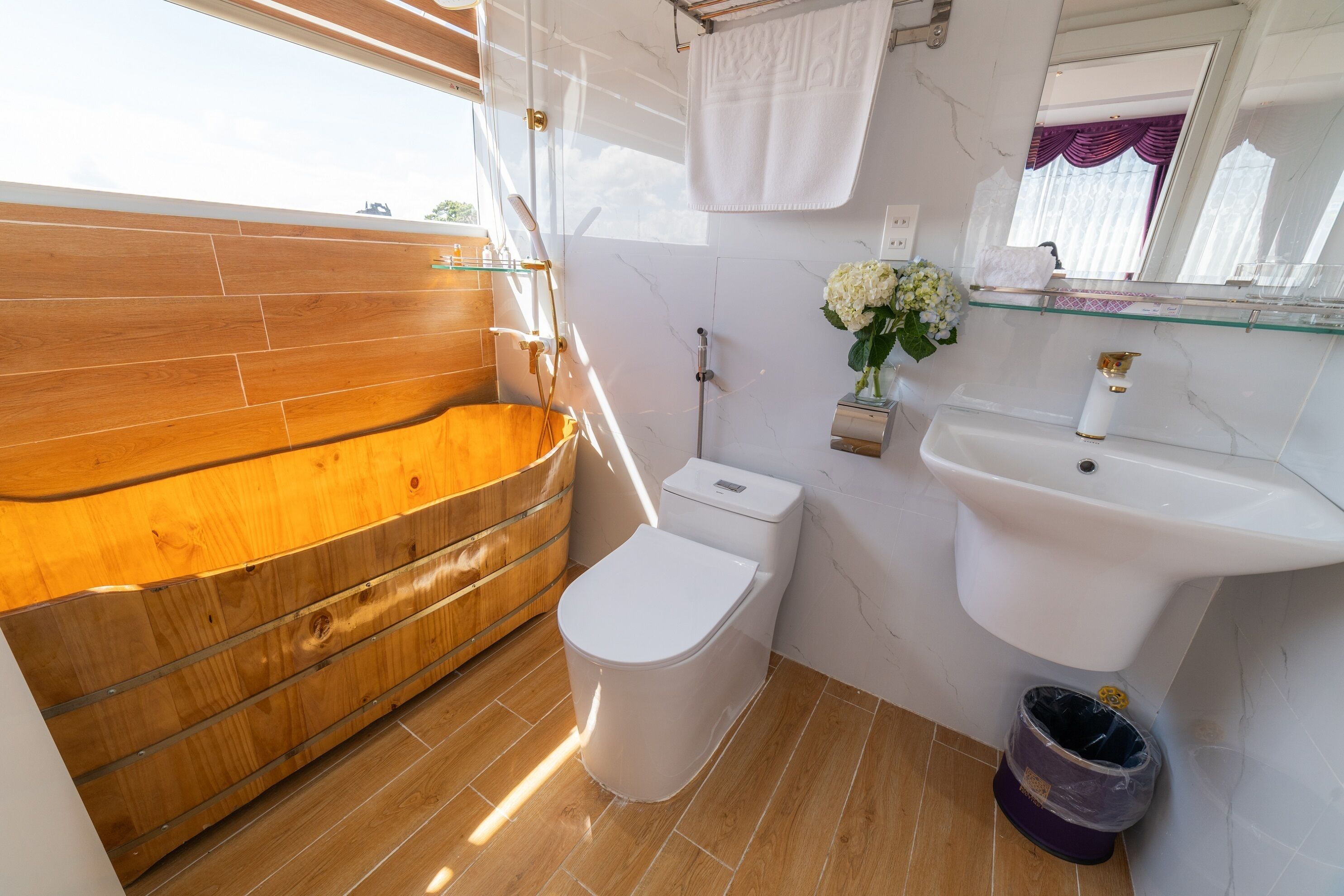 A modern bathroom with a wooden bathtub, a white toilet, a glass shelf with toiletries, and a marble sink.